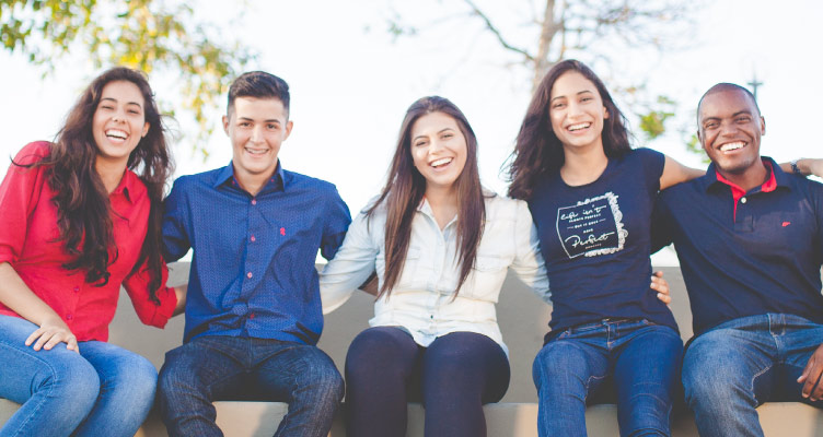 Group of 5 smiling dark-haired young adults sitting on a stone bench outside