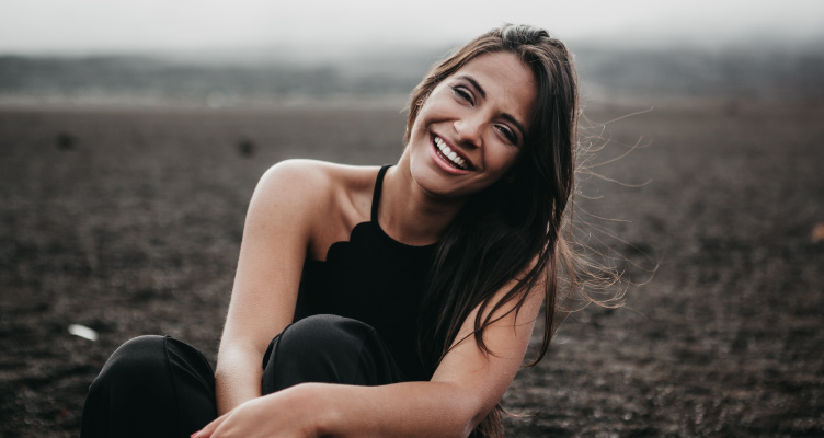 Brunette woman wearing dental veneers smiles while sitting elegantly in a dirt field
