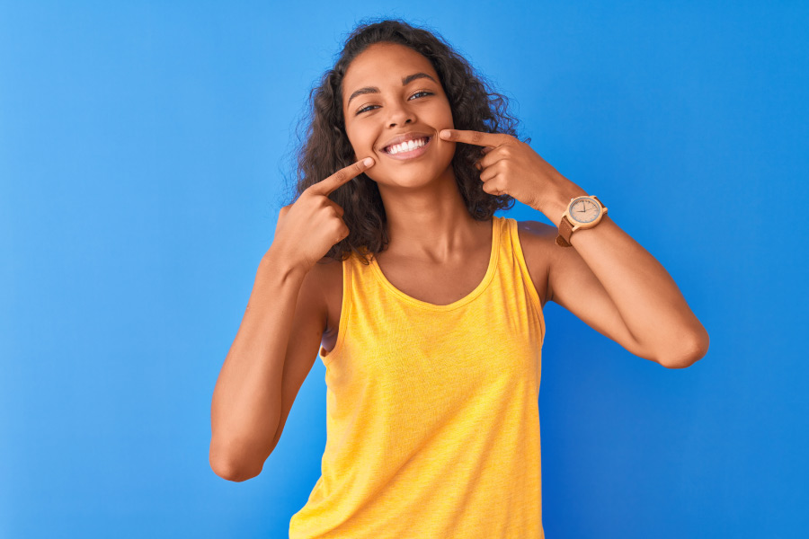 Brunette woman in a yellow tanktop smiles after root canal therapy and points to her teeth