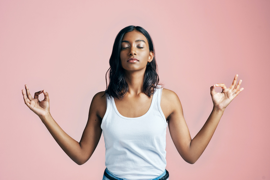 Brown woman in a white tank top and eyes closed stands meditating in a zen pose with her index fingers touching her thumbs