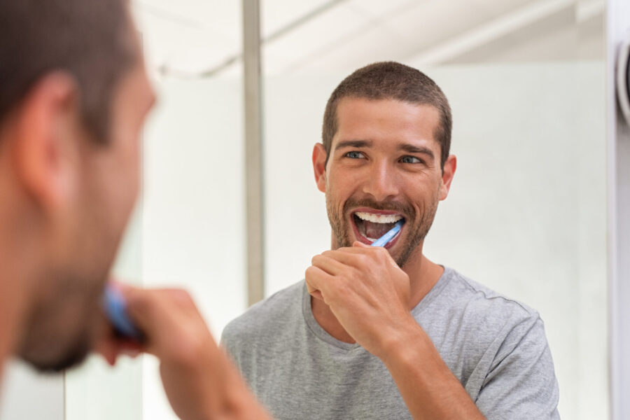 Brunette man in a gray t-shirt smiles in the bathroom mirror as he brushes his gums and teeth
