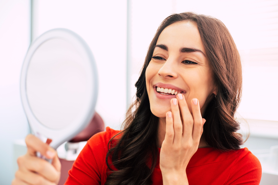 Brunette woman in a red blouse smiles at herself in a hand mirror after a cosmetic makeover at the dentist