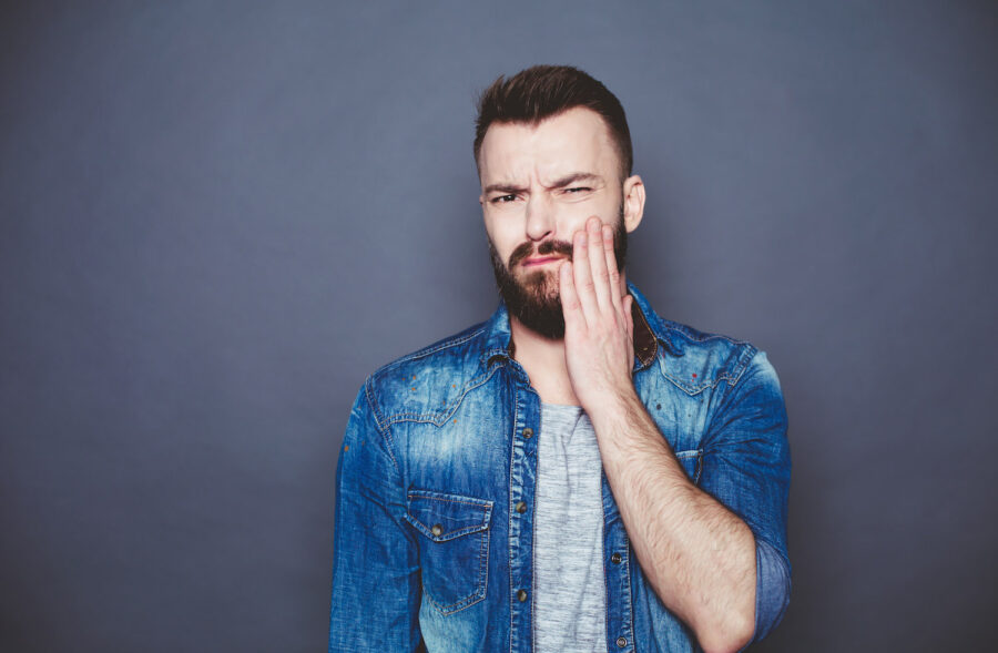 Brunette man in a denim jacket cringes in pain and touches his cheek due to sensitive teeth